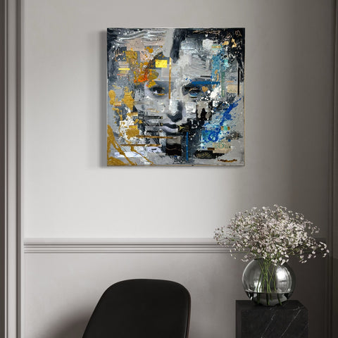 The MIND I by Marjolein Marx displayed in a modern interior. The artwork features a bold, layered portrait with abstract textures in blue, gold, and grey tones, hung above a black chair and a vase with baby's breath flowers.